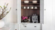 Image of a coffee nook in a built-in cabinet in a white kitchen. There is a silver coffee machine with several matching accessories around it and a vase of pink flowers. There are white mugs on the shelf above the machine.