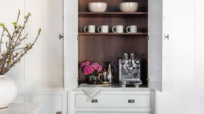 Image of a coffee nook in a built-in cabinet in a white kitchen. There is a silver coffee machine with several matching accessories around it and a vase of pink flowers. There are white mugs on the shelf above the machine.