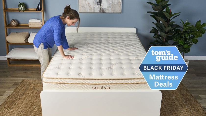 The image shows a dark haired woman wearing a blue shirt and beige trousers placing her hands on the top of a cream and gold mattress, placed on a white bed frame in a neutrally decorated bedroom. On the top of the image a badge reads 'Tom's Guide Black Friday Mattress Deals' 