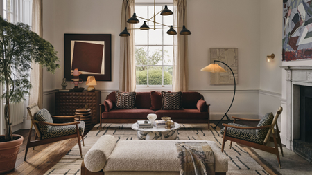 A layered lighting scheme in a modern living space with a high ceiling, a dark red-brown sofa at the far wall in front of a large window with floor-length curtains, facing a daybed, with two armchairs on either side, and artwork adorns the cream walls alongside a large potted plant and a fireplace