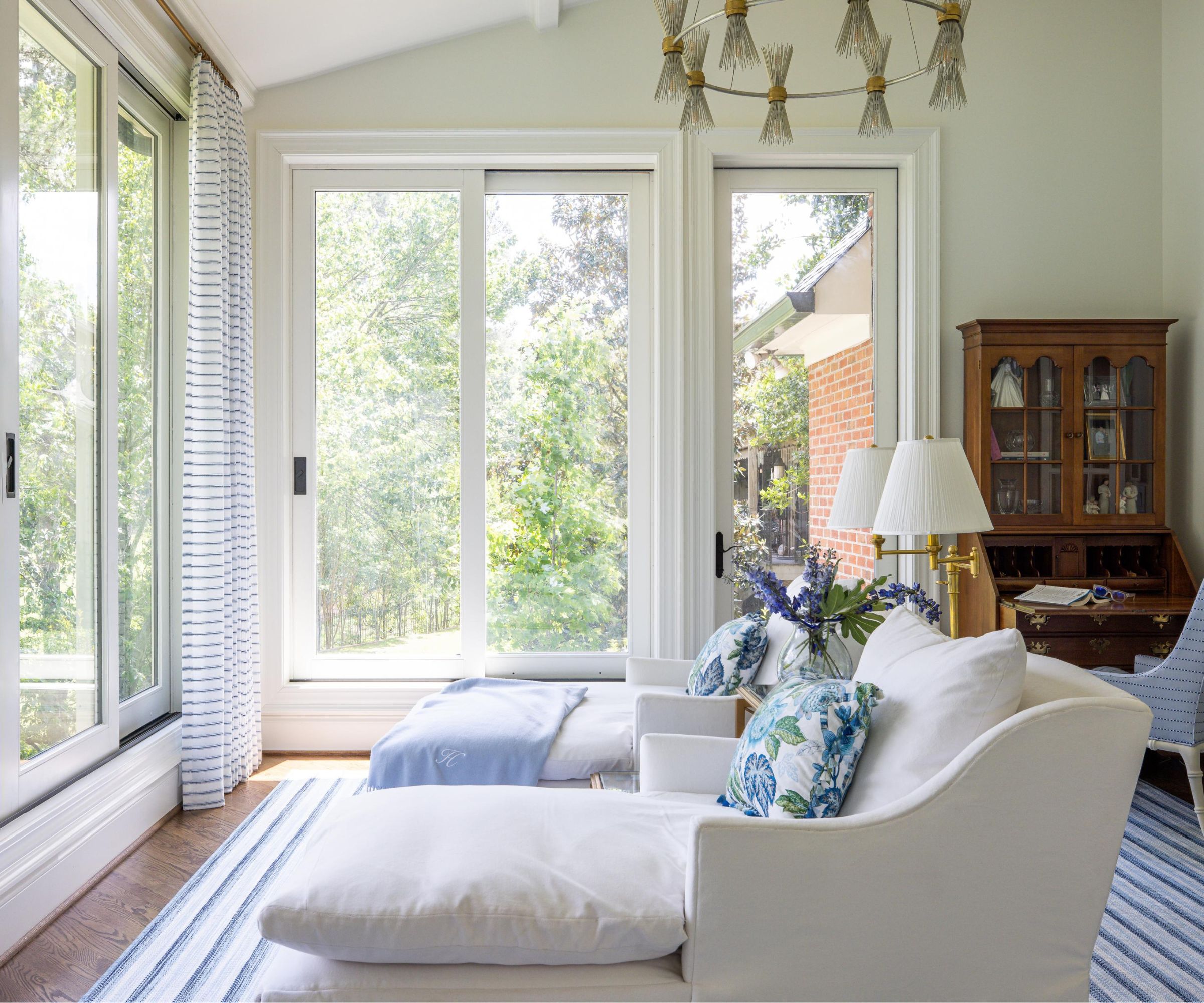 Sunroom with two large sliding doors that open onto a terrace, two cream linen lounge armchairs with floral cushions, a blue striped rug, two gold pleated lamps and a mahogany desk in the background.