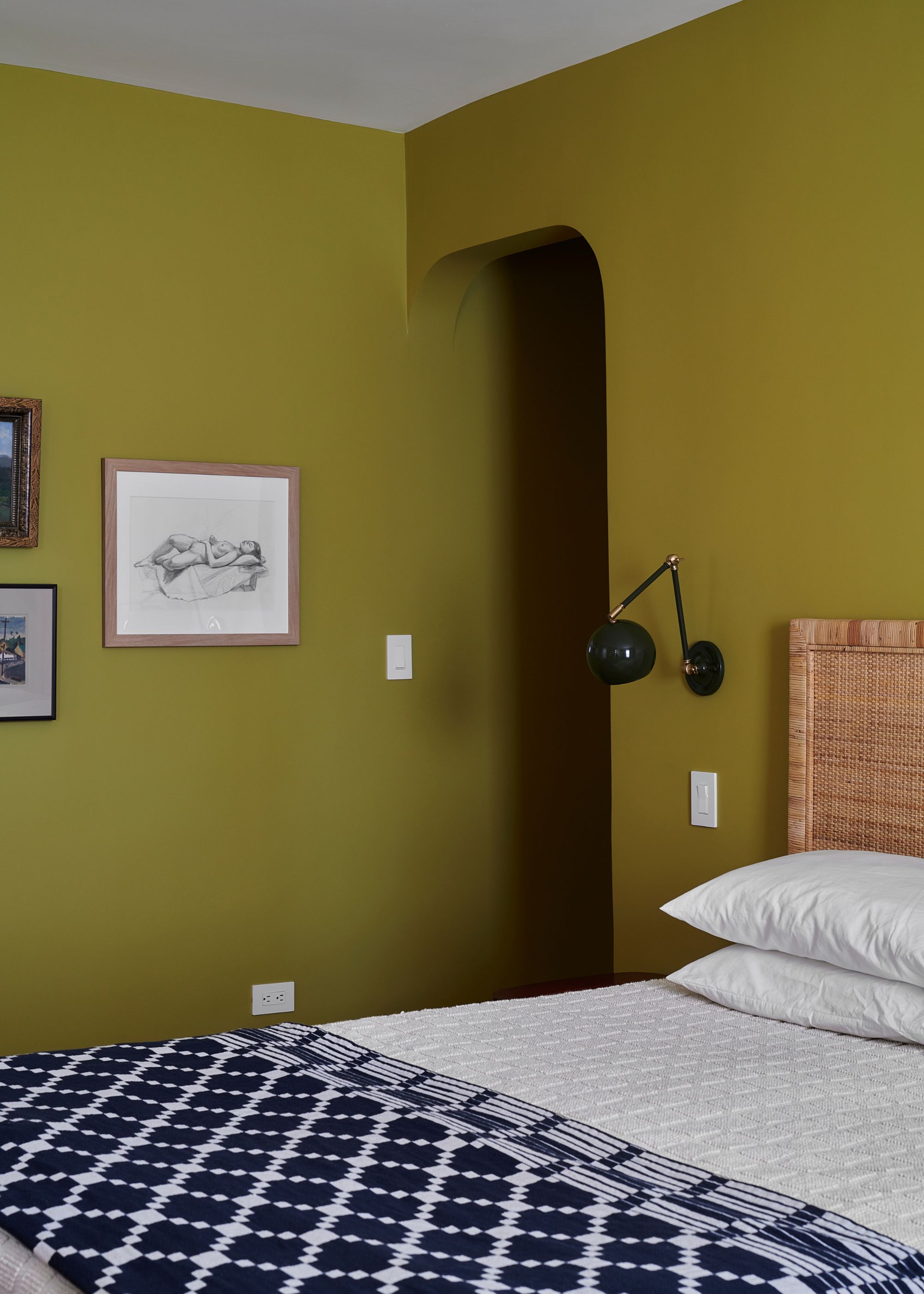 A small bedroom with dark chartreuse walls, a white ceiling, a rattan headboard with white bedding and a navy and white patterned quilt, a small gallery wall, and a black wall light.