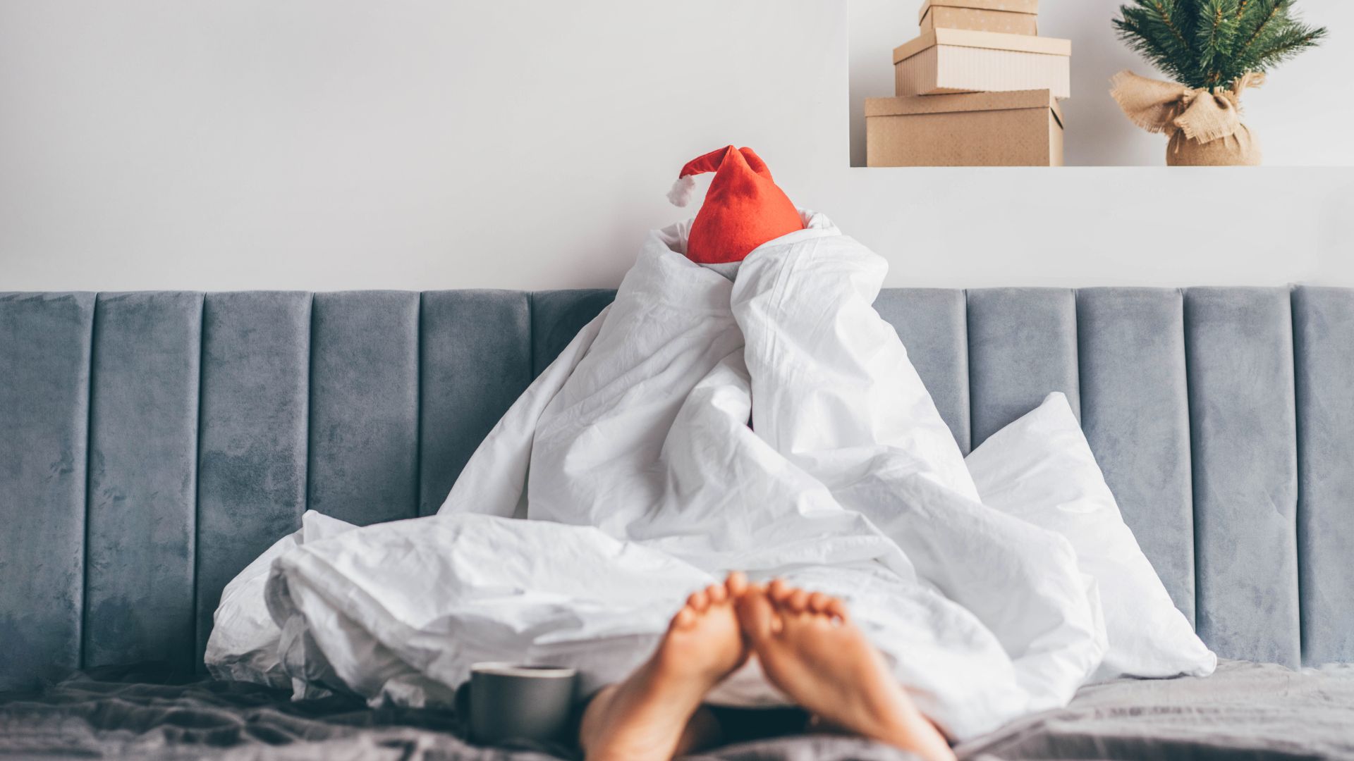 A person sitting in bed wrapped up in a white duvet that covers their whole body except their feet. A Santa hat is visible poking out of the top of the duvet.