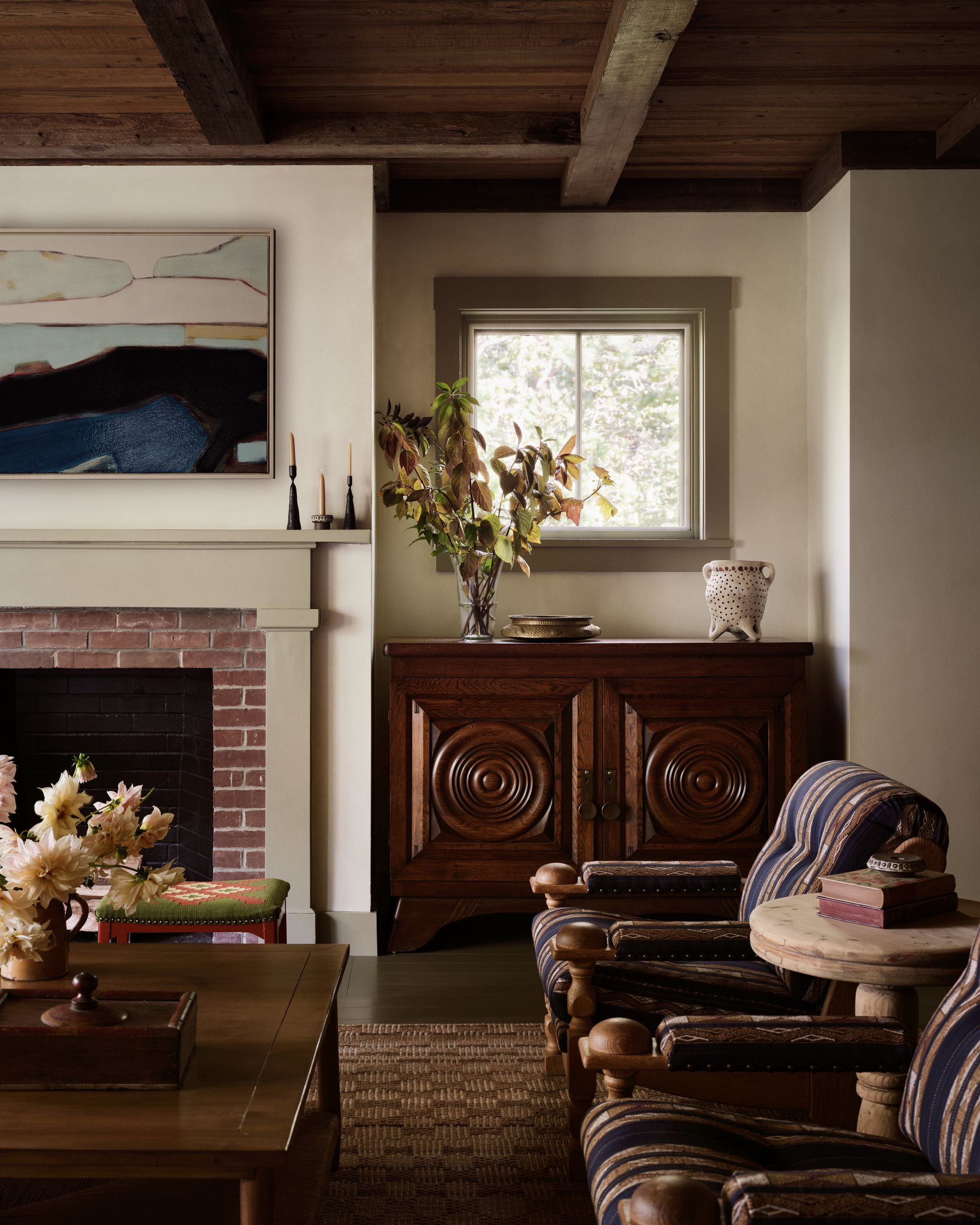 Image of a sitting area in a traditional living room. There is a large dark wood cabinet, two blue striped accent chairs, a brick fire place, a wooden coffee table, and art on the walls.