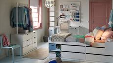 An IKEA dorm room with a white raised bed with colorful bedding, white chest of drawers, white clothes rack, blue chair, white desk, pink door, and pink framed window filling the room with light.