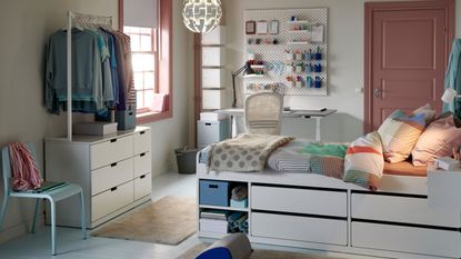 An IKEA dorm room with a white raised bed with colorful bedding, white chest of drawers, white clothes rack, blue chair, white desk, pink door, and pink framed window filling the room with light.