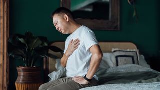 man suffering from chest pain as he sits on his bed