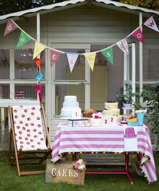 A table of cakes set up in front of a summer house with a deckchair and traditional bunting