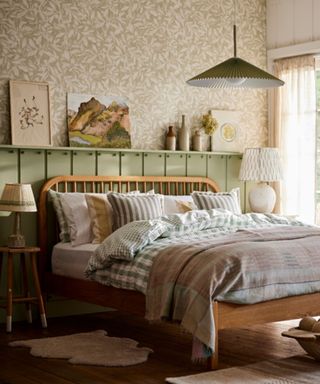 A bedroom with brown and white leafy wallpaper, green paneling, a wooden bed with gingham bedding, and a wooden floor with white rugs on it