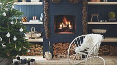 A dark blue living room with panelled walls, shelves, and a brick fireplace with a lit fire. Around the fire is a pinecone garland, and to the left in the foreground is a large Christmas tree decorated with white ornaments. To the right of it is a large metal white statement chair with white fluffy cushion.