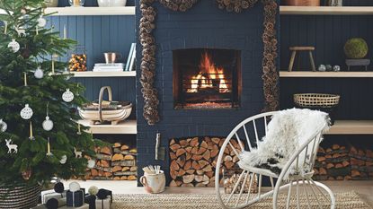 A dark blue living room with panelled walls, shelves, and a brick fireplace with a lit fire. Around the fire is a pinecone garland, and to the left in the foreground is a large Christmas tree decorated with white ornaments. To the right of it is a large metal white statement chair with white fluffy cushion.