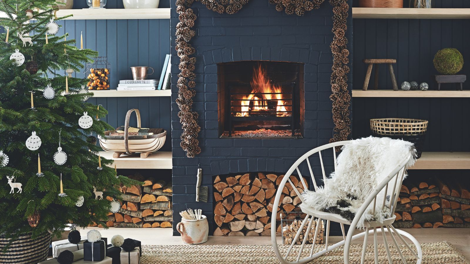 A dark blue living room with panelled walls, shelves, and a brick fireplace with a lit fire. Around the fire is a pinecone garland, and to the left in the foreground is a large Christmas tree decorated with white ornaments. To the right of it is a large metal white statement chair with white fluffy cushion.