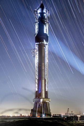 A large stainless steel rocket stands on the launchpad. the finned top half is covered in heat tiles. The streaks of stars from long camera exposures line the night sky.