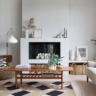 neutral living room with alcoves in the wall containing candles on the top one and a wood log pile at the bottom, a grey, brown and black pattern rug, with a brown coffee table on top
