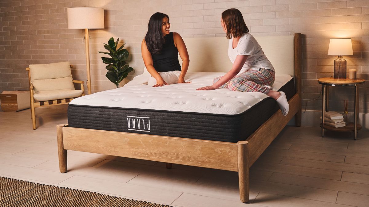 Two people sit on top of the Plank Firm flippable double-sided mattress in a bedroom