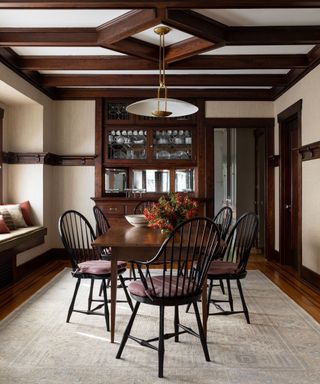 Dining room with dark wood beams, paneling and features. Dark wood dining table with four black wood dining chairs, large cream rug, window seat with cushions