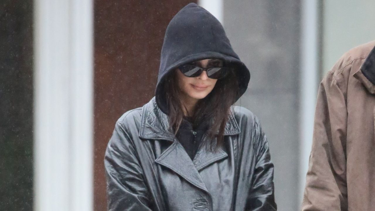 Emily Ratajkowski walks in the rain wearing a black hoodie and leather jacket