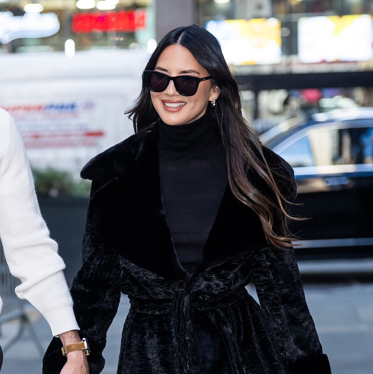 Olivia Munn with John Mulaney, wearing a black coat and turtleneck, stovepipe blue jeans, and black pumps