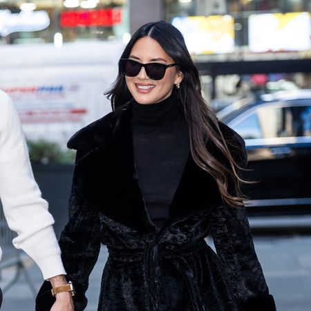 Olivia Munn with John Mulaney, wearing a black coat and turtleneck, stovepipe blue jeans, and black pumps