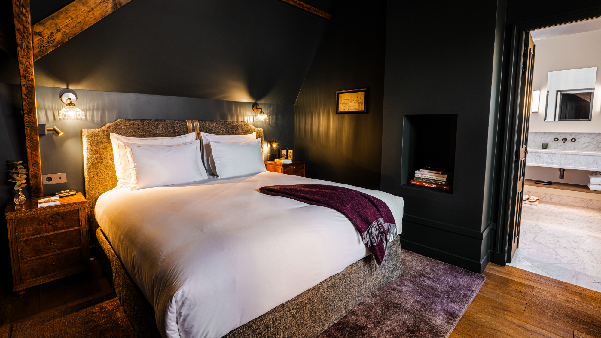 King size bed with throw, dark blue walls and wooden beams, with view through doorway to a bathroom