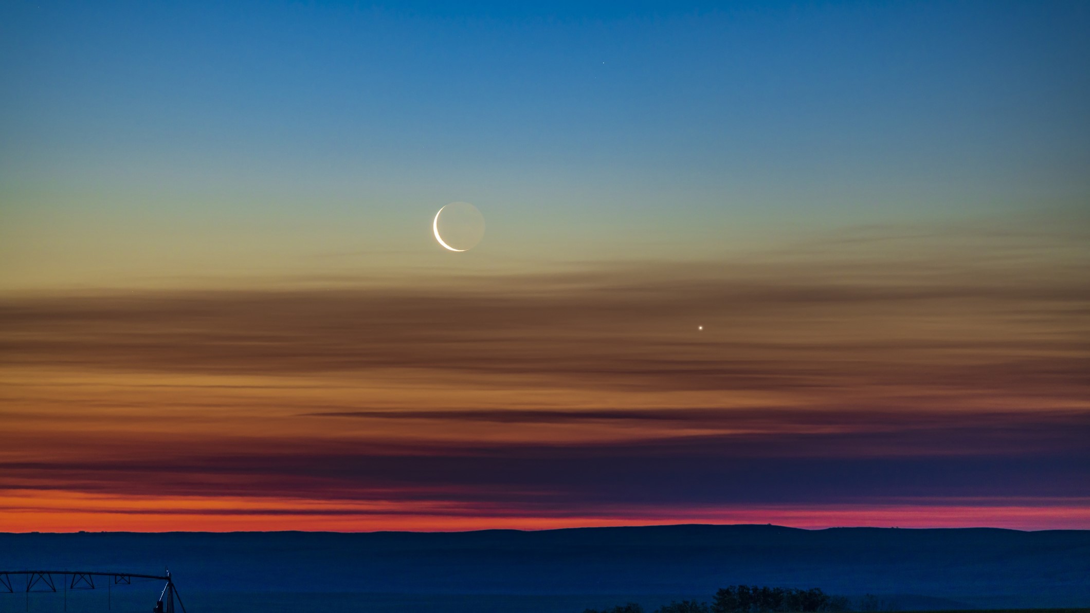 Die Konjunktion der abnehmenden Sichel und der Venus, wie sie am nordöstlichen Morgenhimmel im Süden von Alberta, Kanada, aufgehen. Das Licht der Erde ist auf der dunklen Seite des Mondes zu sehen. Der Himmel zeigt einen wunderbaren Farbübergang von Orange am Horizont über das gesamte Spektrum bis hin zu Blau am oberen Ende