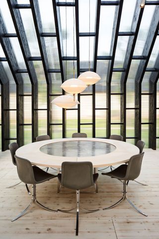 Inside image of the pavilion, wood structure, slanted glazed roof, round dining table with eight beige chairs, cream oval ceiling lights suspended over the table, natural light wood floor
