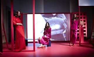 three female models on a set, two standing and one sitting down, one is wearing red and the others are wearing pink