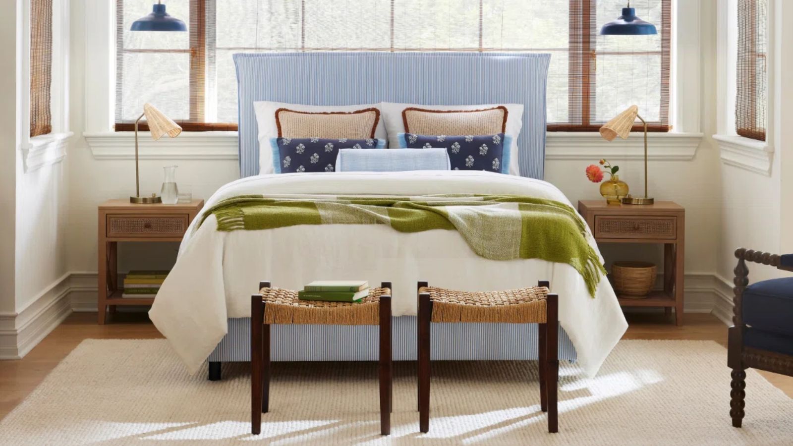 wayfair styled bedroom with a blue and white striped bed wooden nightstands, little wooden and wicker stools at the foot of the bed and green blanket on the end