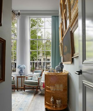 A light blue room with floor-ot-ceiling windows, a rolling bar, and a collage of vintage gold picture frames