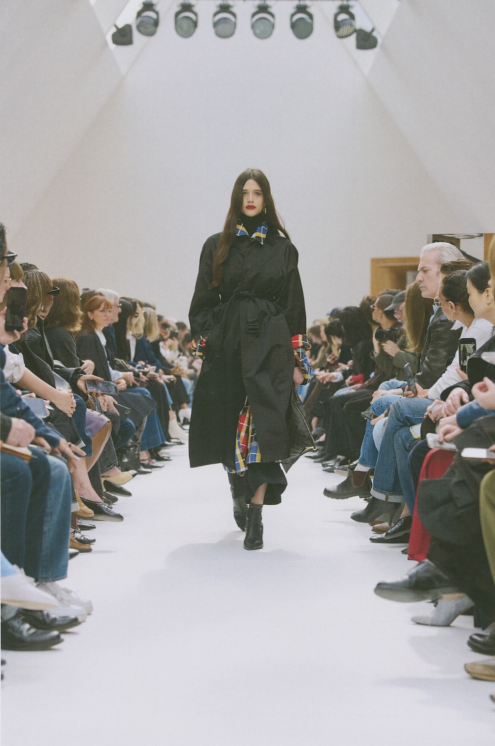 a model on the Celine runway wearing a black trench coat with plaid layers underneath
