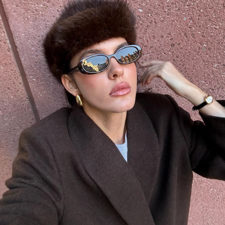 Fashion influencer @sobalera wearing a rich-looking winter outfit with a brown wool coat, faux fur hat, oval sunglasses, gold earrings, and an elegant leather-strap watch.