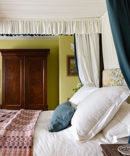Bedroom with citron green walls, a four-poster bed with a blue and white lined canopy on top, a dark mahogany wardrobe and white bedding with a pink Welsh blanket on top.