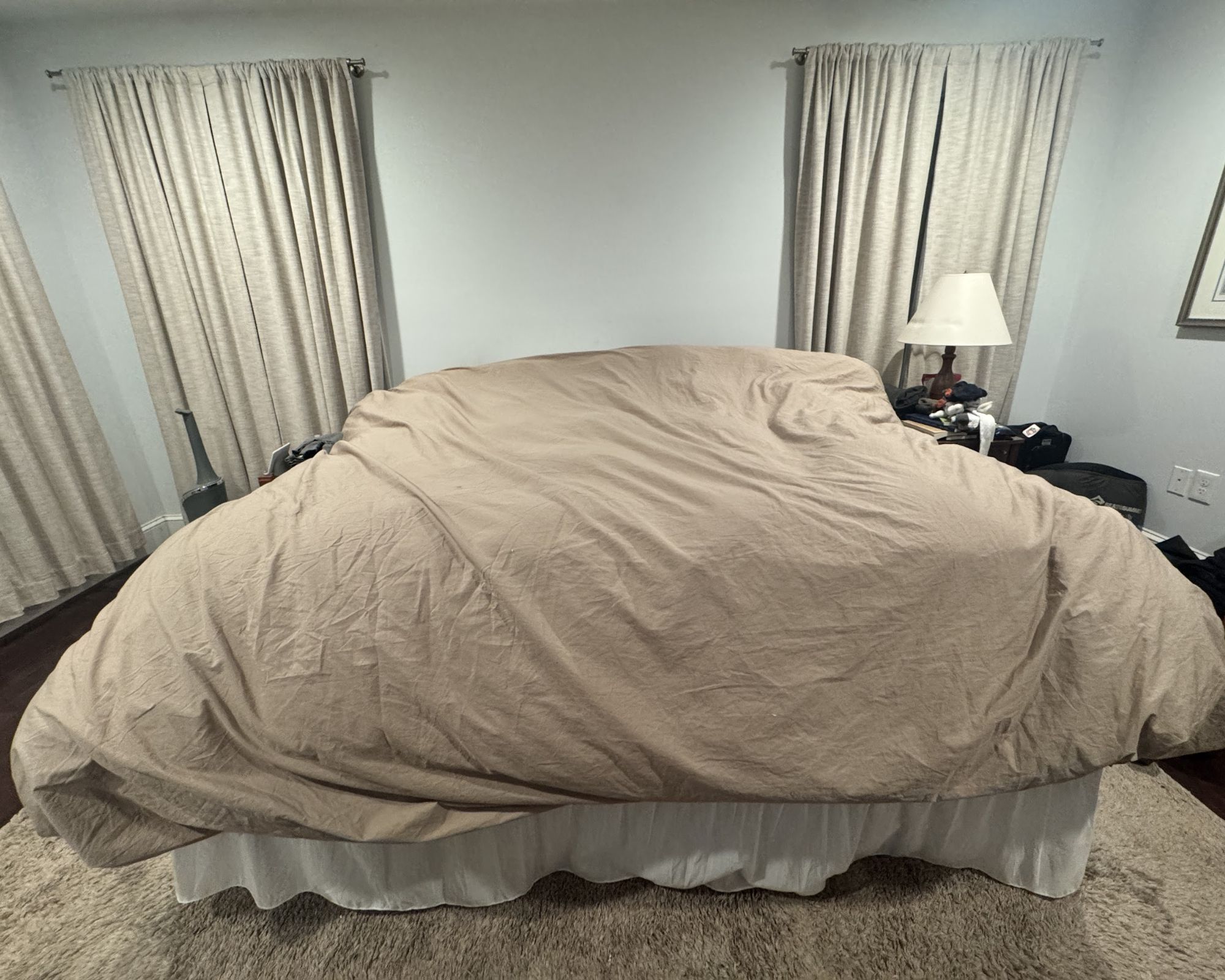 A bed with a white bed skirt and tan comforter in a room painted blue with tan curtains and a tan rug.