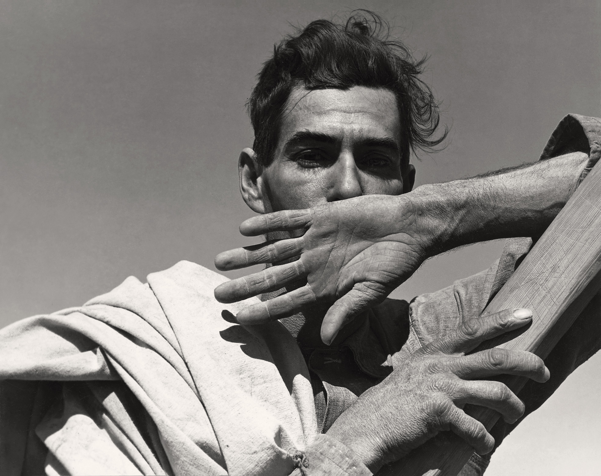 A migratory cotton picker rests his large, deeply lined hands across his face and looks directly into the camera against a plain sky, in a formally composed close-up portrait shot in Arizona.
