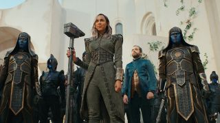 Dar-Benn (shoulder length gray/light purple hair and wearing a grey outfit) is holding a large hammer/staff whilst standing in front of an army of Kree (blue humanoids).