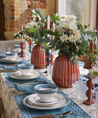 Close up of decorated dining table with orange, blue and cream patterned tablecloth, blue fabric place mats, white and blue plates and bowls laid on table, orange vases with flowers and greenery, orange candleholders with orange, cream and pink candles