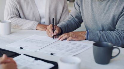 An older man signs a document at a table, only his hands and torso showing.