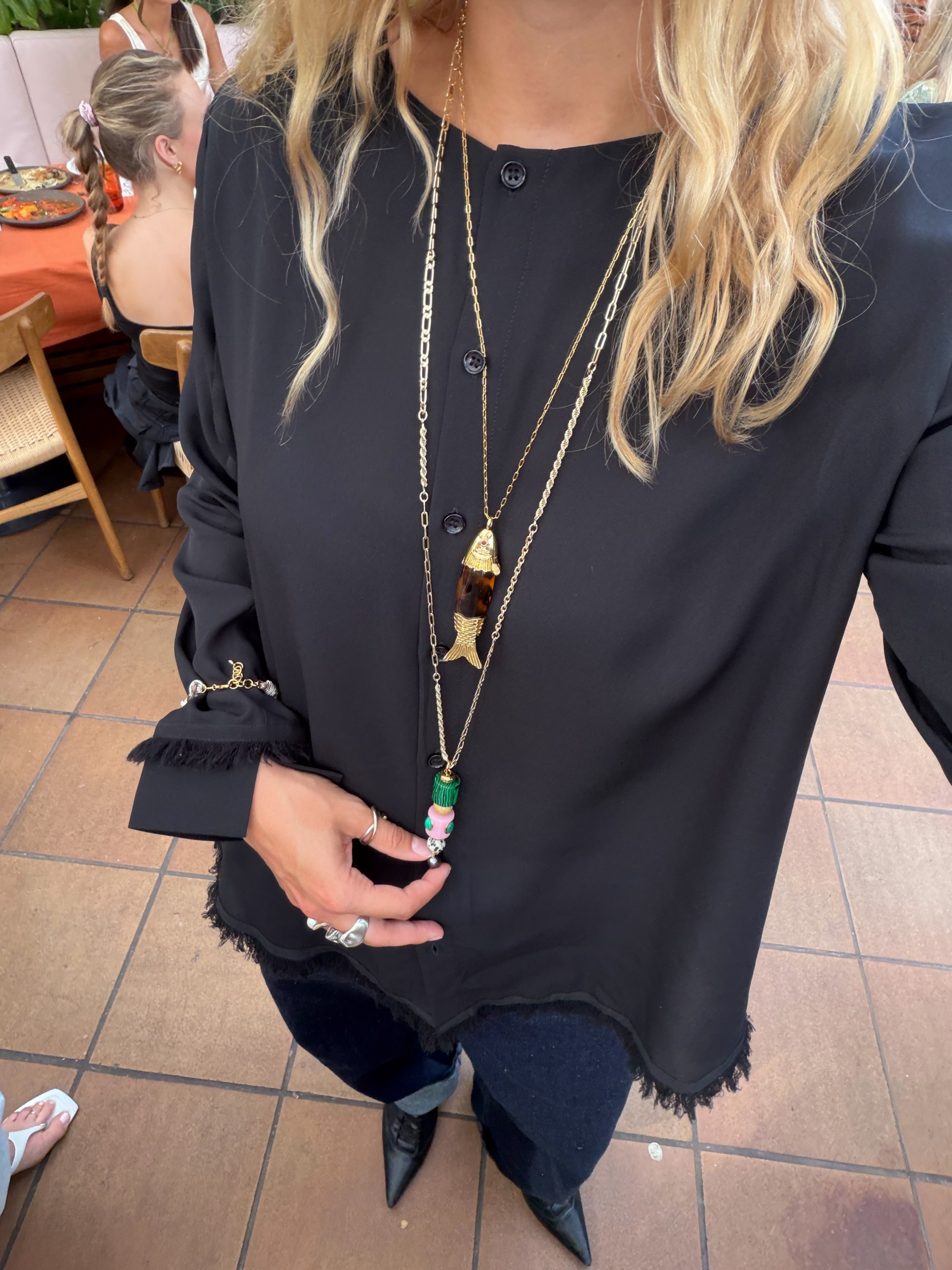 An image of an influencer wearing a layered necklaces over a black, long-sleeve top, dark indigo jeans and black lace-up heels
