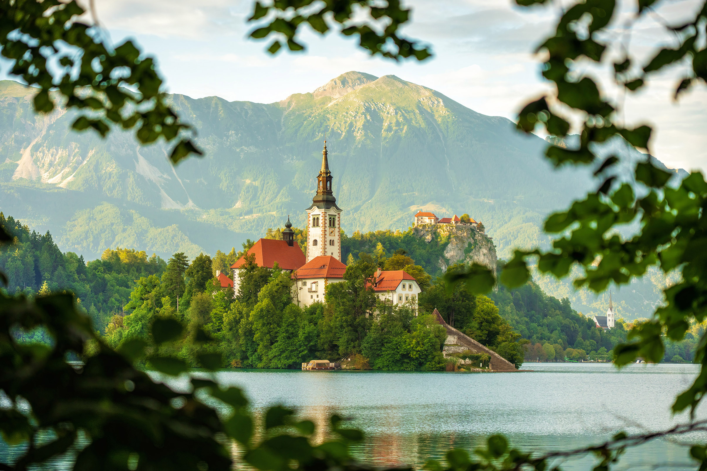A picturesque island on a serene lake features a church with a tall bell tower, framed by lush green leaves. Majestic mountains rise in the background