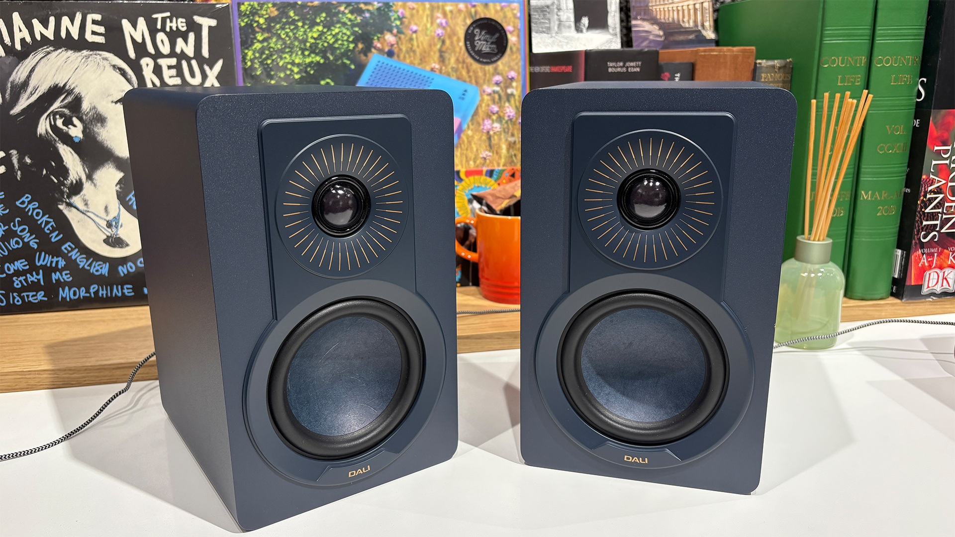 Dali Kupid standmount speakers on white wooden surface in front of busy bookcase
