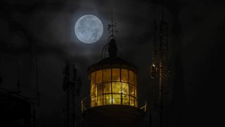 a dark and moody image showing a dimly lit lighthouse glowing a faint yellow color and a moon shining brightly in the distance behind thin clouds.
