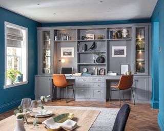 shaker style home office, with built-in grey units with two small desk areas and lots of shelves with books, art and vases on it, and a dining table in the foreground of the picture