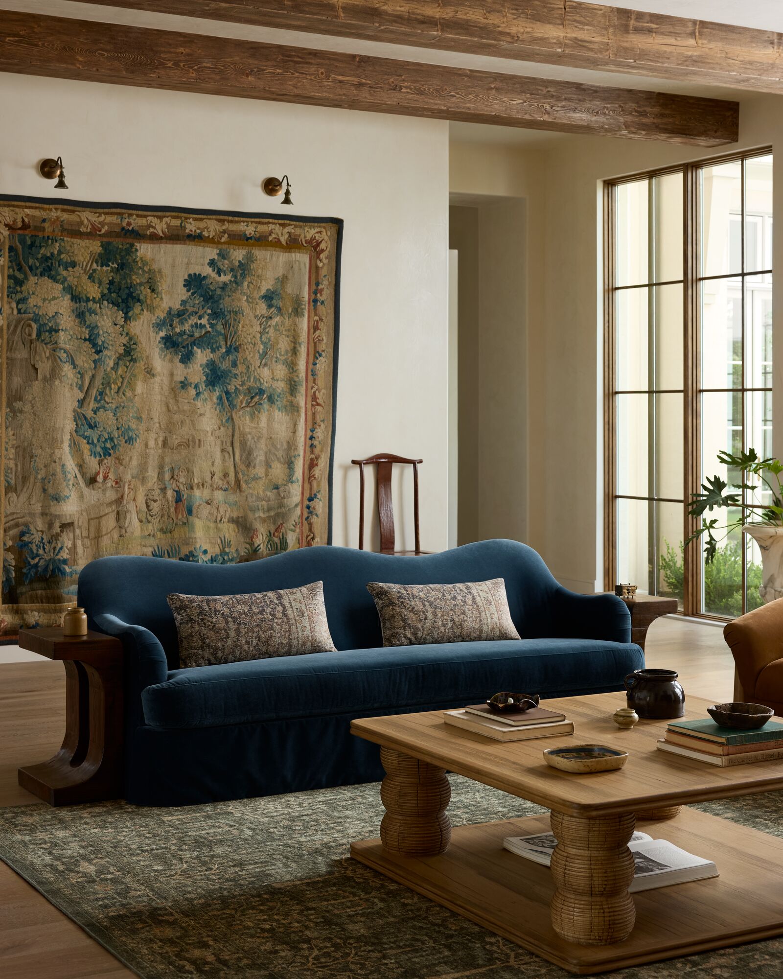 Peacock blue scalloped edge velvet sofa and a wooden coffee table anchored by a green patterned rug and tapestry hanging in the back