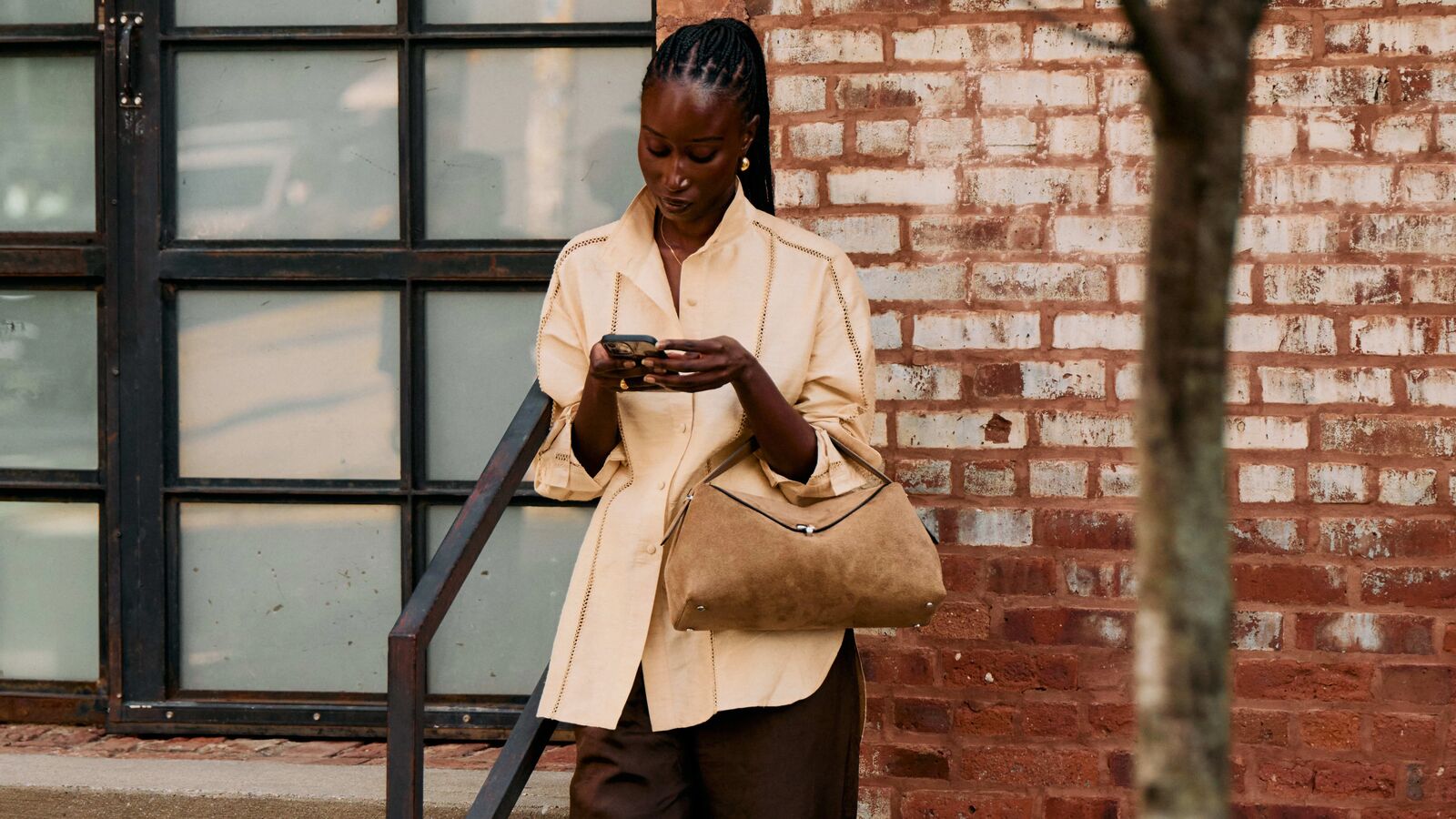 Woman wearing a beige button-down with brown pleated trousers and a suede bag looking at her phone.