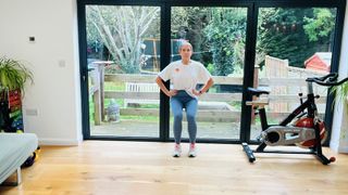 Trainer Maddy Biddulph performs a wall sit in her living room. Her knees are bent and her back rests against a glass door. Behind her we can see a garden and next to her is an exercise bike.