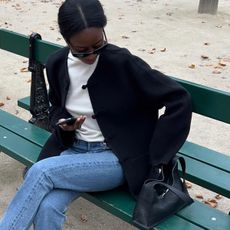  @sylviemus_ wearing a cream sweater, black cardigan, and jeans while sitting on a Paris park bench.