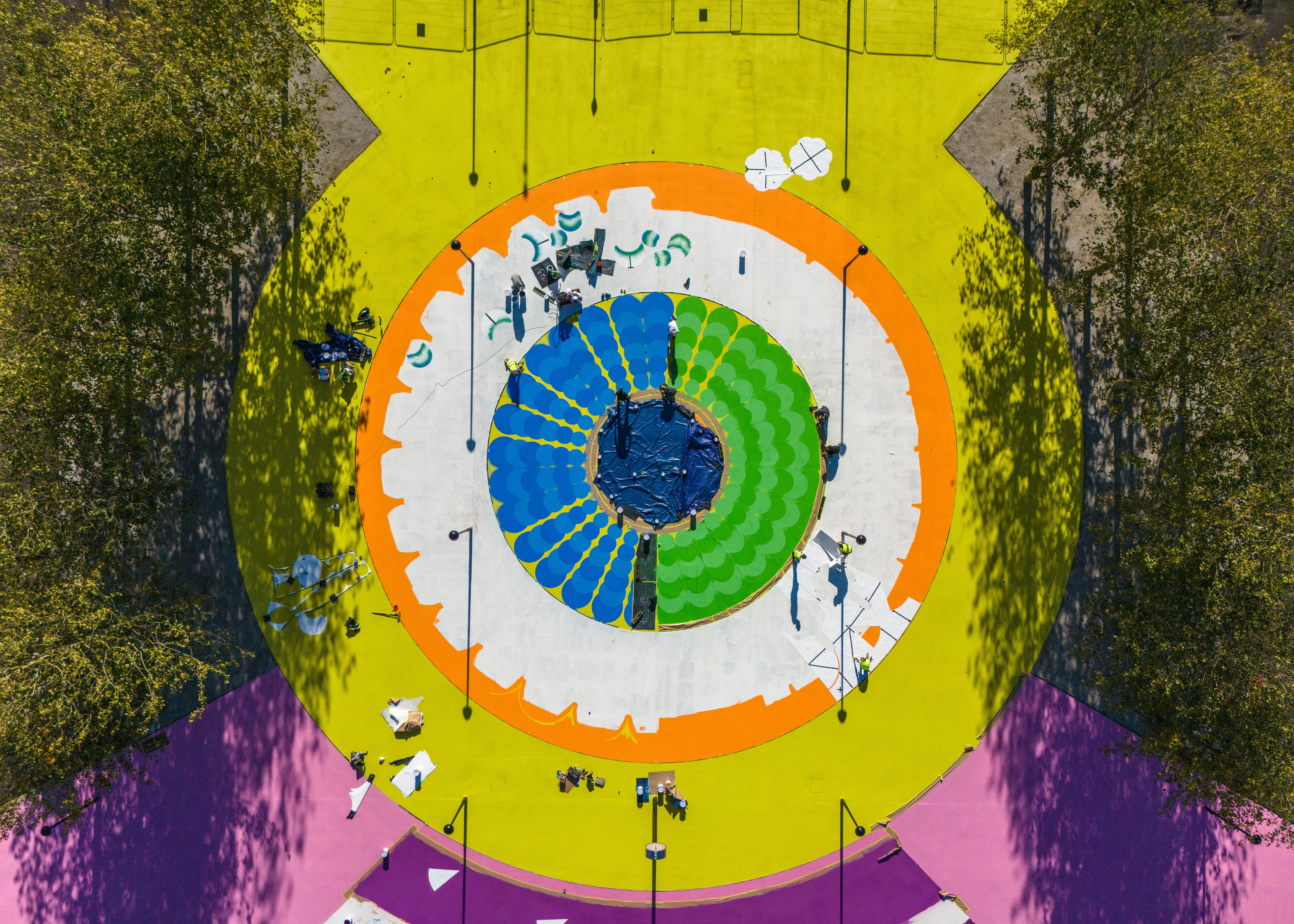 A colorful, concentric mural in yellow, orange, green, and blue, captured in work in progress stages from high above.