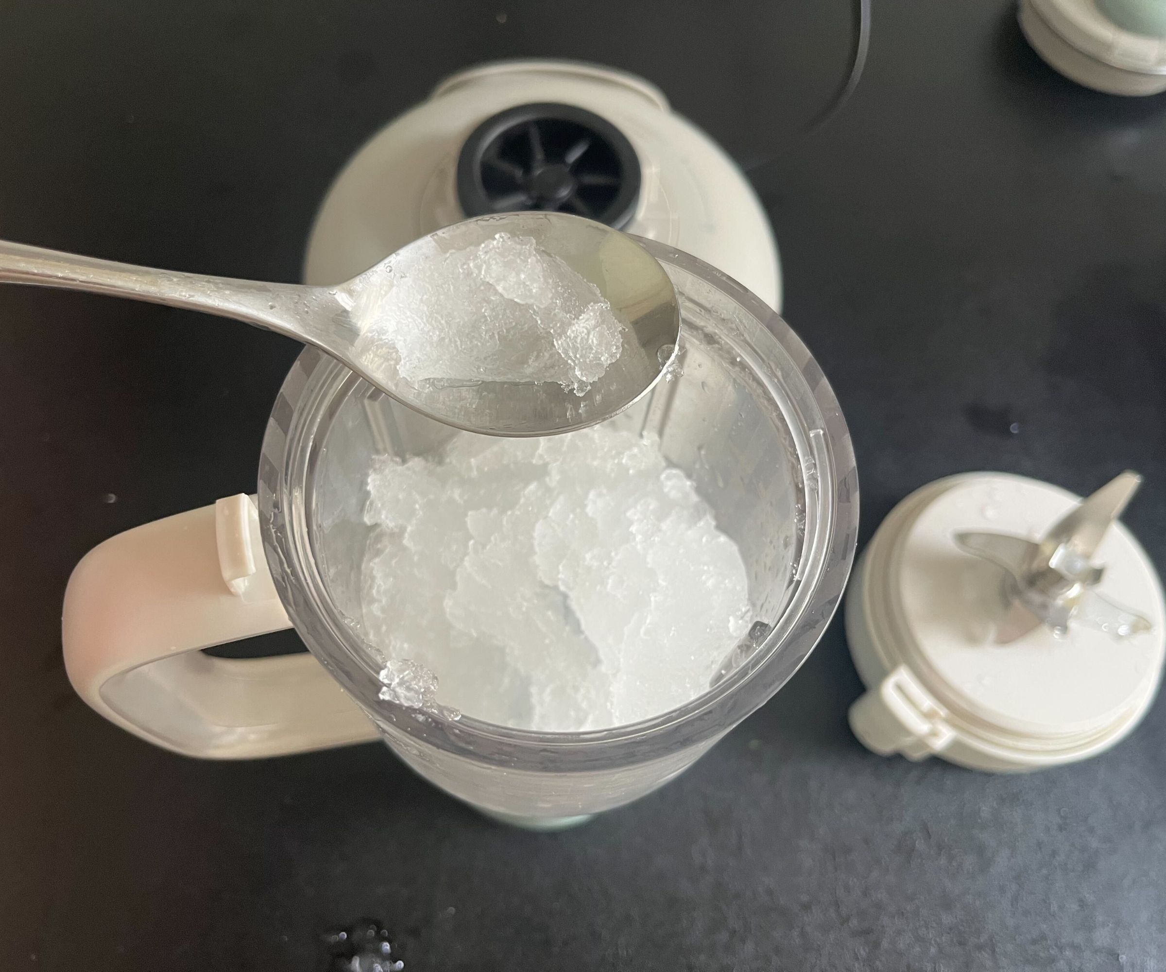 A large chunk of ice on a spoon above a tumbler of crushed ice on a black counter