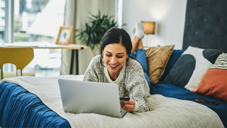 A woman lying on her stomach in bed on her laptop, smiling holding a credit card