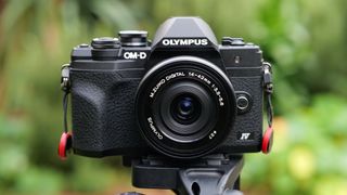 Kameraet Olympus OM-D E-M10 Mark IV på et stativ, med uskarpt bladverk i bakgrunnen.